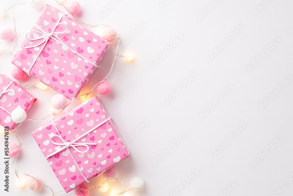 Valentine's Day concept. Top view photo of pink gift boxes in wrapping paper with heart pattern light bulb garland and fluffy pompons on isolated white background with empty space