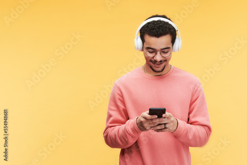 Happy guy in headphones, eyeglasses and pink sweatshirt scrolling through playlist or online audio files in smartphone in isolation © pressmaster