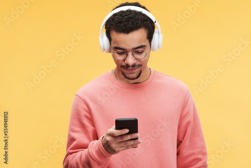 Young man in pink pullover listening to music in headphones and choosing new sound tracks in mobile phone in front of camera © pressmaster