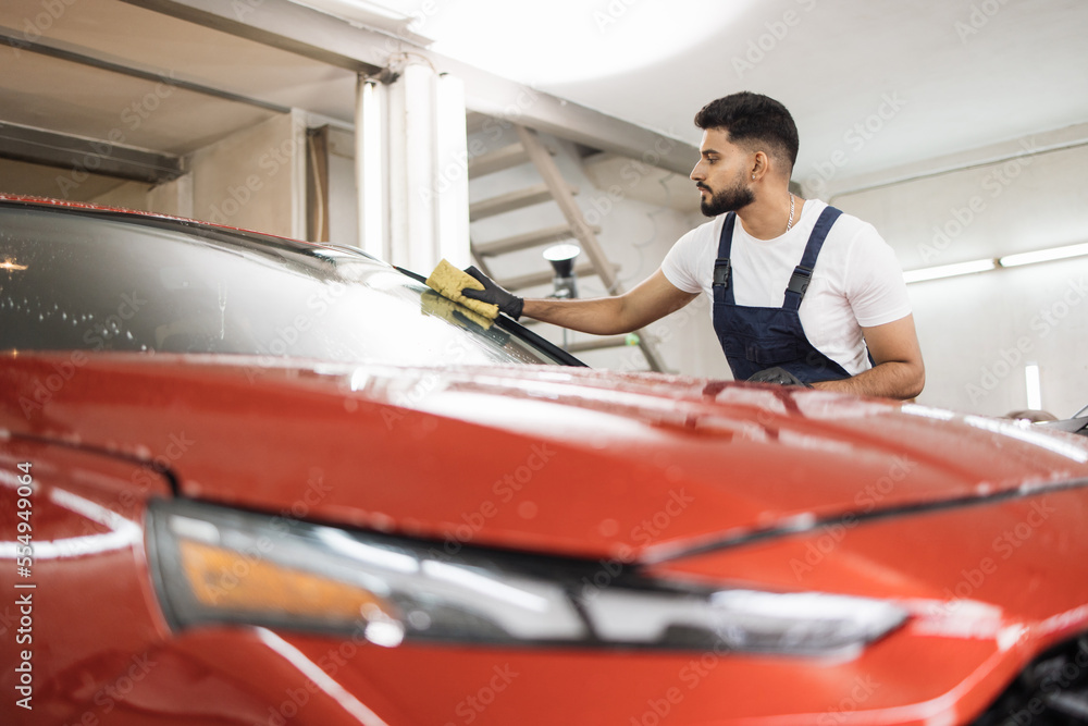 Fototapeta premium Confident young man, car wash worker, wearing t-shirt and overalls cleaning the windshield of car with the help of special foam and sponge. Washing the car by hand, sponge and foam bubbles.
