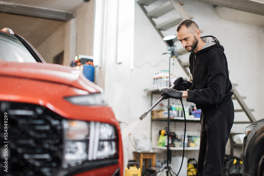 Young good-looking bearded man worker washing luxury red crossover car on a car wash, holding high pressure water washer. Car wash station concept.