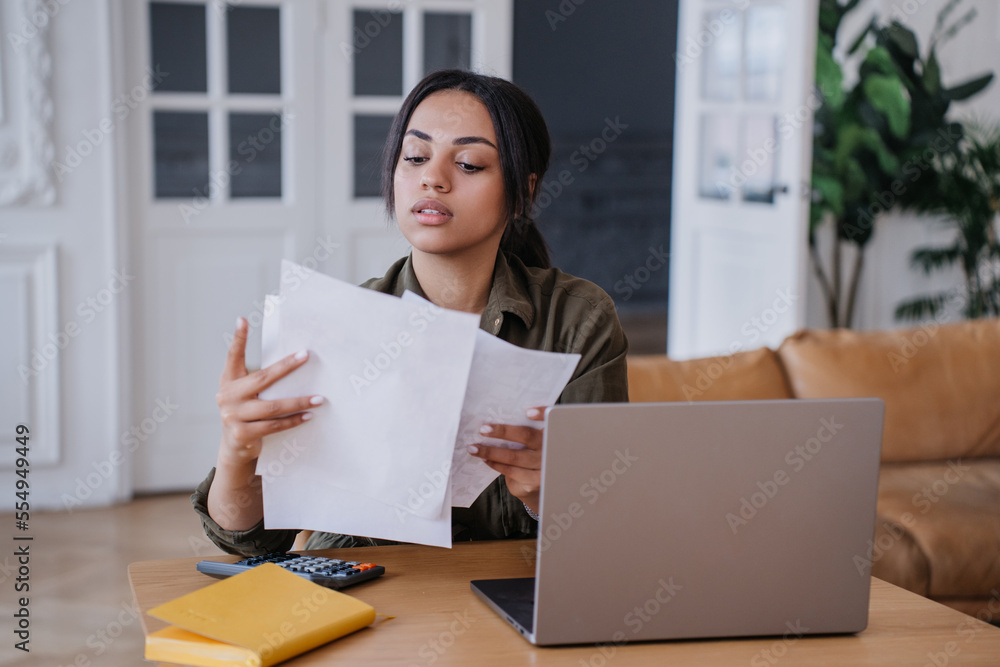 Thoughtful confident Brazilian young woman sitting at table with laptop and calculator, holding paper sheets. Attractive African American businesswoman thinking about new project. Business people. Thoughtful confident Brazilian young woman sitting at table with laptop and calculator, holding paper sheets. Attractive African American businesswoman thinking about new project. Business people.