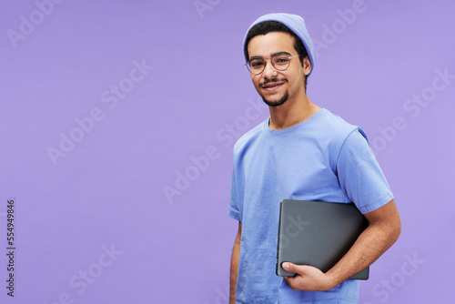 Young smiling businessman or student in blue t-shirt and beanie holding laptop while standing on violet background and looking at camera