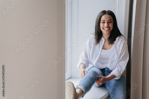 Cheerful young African American female in casual dress looks at camera sitting on windowsill laughing looks aside. Romance, people in love. Pretty Brazilian girl having fun indoors