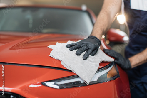Cropped image of hand of male professional car wash worker in black rubber gloves, holding the gray microfiber and polishing the car hood and headlight of luxury red car.