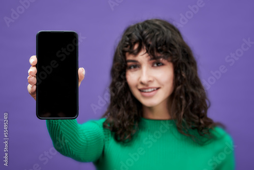 Selective focus on blank screen of smartphone held by young brunette woman in green pullover standing against violet background