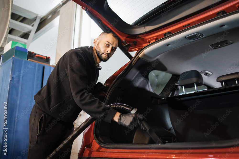 Young confident car wash service worker, doing wet cleaning of luxurious vehicle luggage trunk using vacuum cleaner. Car detailing concept. Young confident car wash service worker, doing wet cleaning of luxurious vehicle luggage trunk using vacuum cleaner. Car detailing concept.