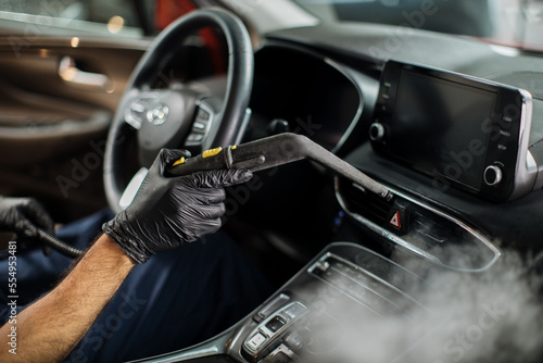 Close up of hands of man in black protective rubber gloves cleaning interior of the car with hot steam cleaner. Selective focus on guy hands. Auto cleaning service and detailing concept. © sofiko14
