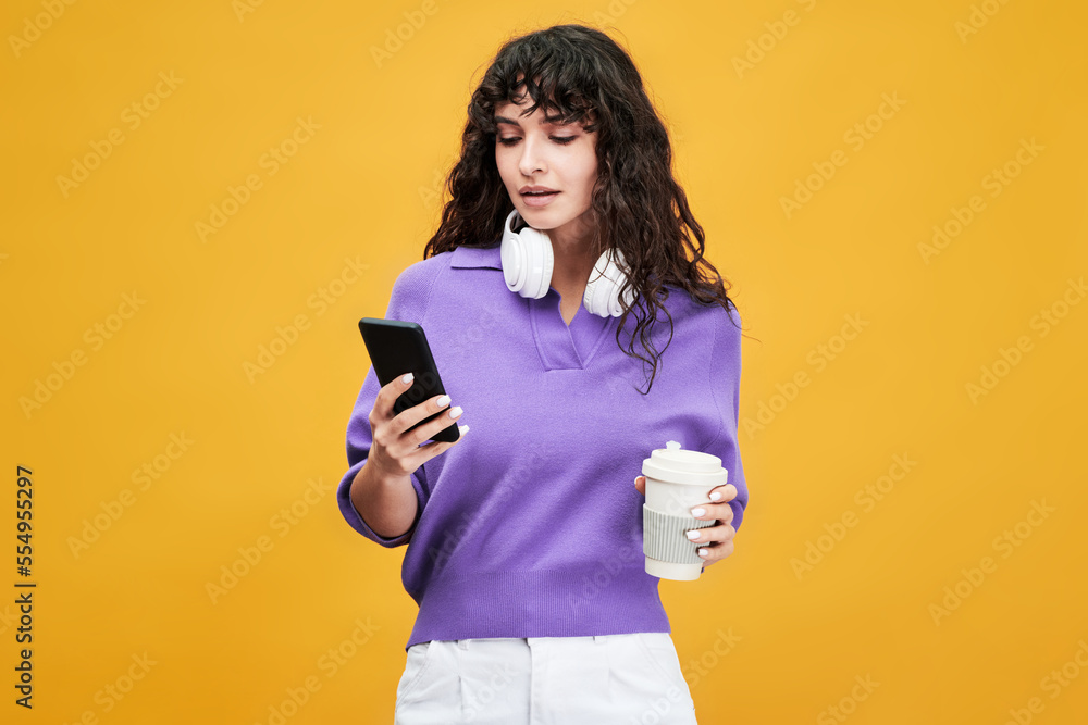 Cute female student in lavender v-shirt and while pants using mobile phone and having cup of tea at break against yellow background Cute female student in lavender v-shirt and while pants using mobile phone and having cup of tea at break against yellow background