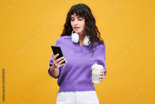 Cute female student in lavender v-shirt and while pants using mobile phone and having cup of tea at break against yellow background