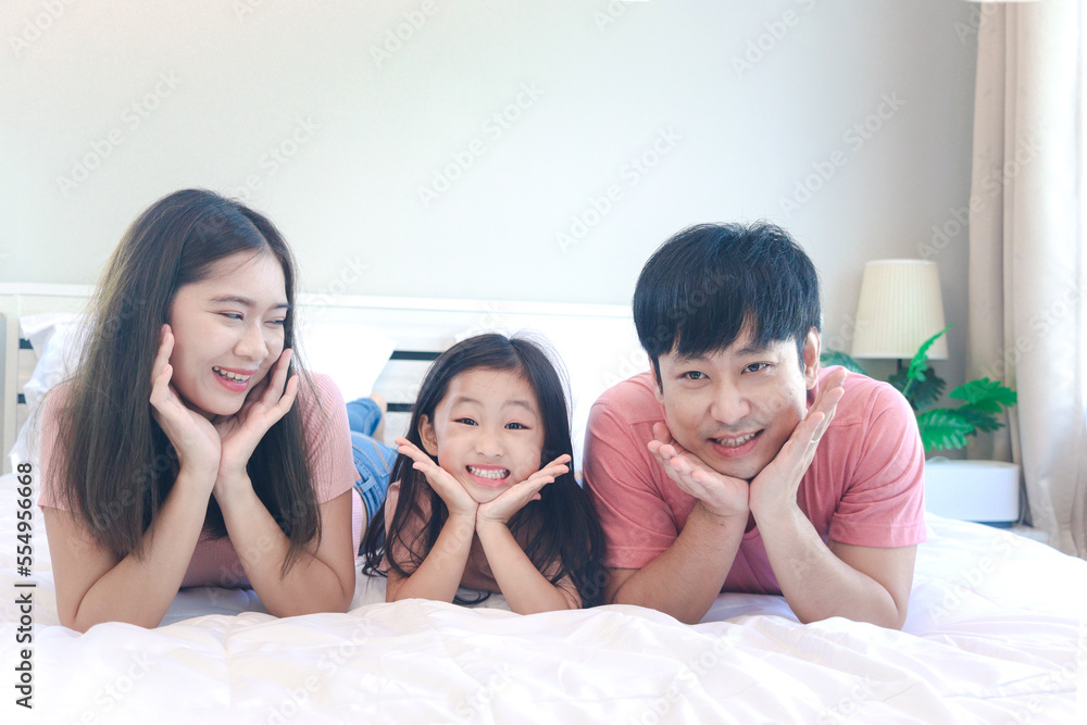 Happy Asian family has fun in bedroom. Father, mother and daughter girl lying on bed together, propping chin up with hands and smiling to camera, dad mom, parents having good memory with kid Happy Asian family has fun in bedroom. Father, mother and daughter girl lying on bed together, propping chin up with hands and smiling to camera, dad mom, parents having good memory with kid