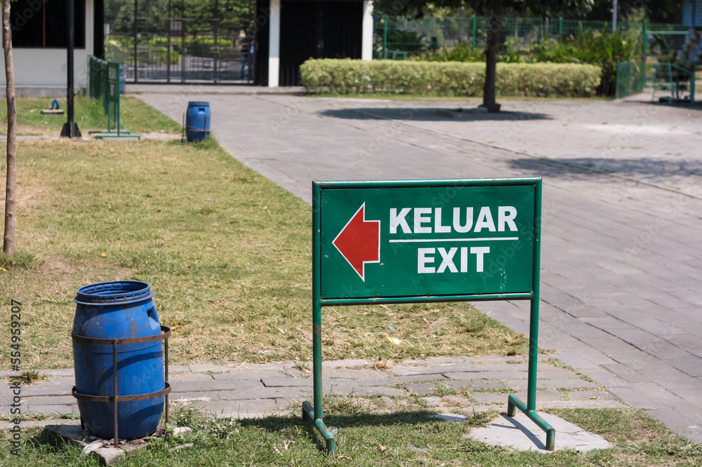View of green metal information board with keluar (exit) red direction arrow in public park with blue plastic trash bin and grass and trees. No people.