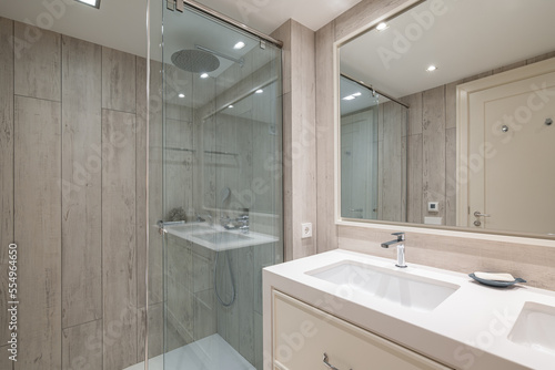 Spacious bathroom with bright artificial lighting  large mirror reflecting front door and pair of snow-white sinks built into light beige cabinet. Shower enclosure with blue glass splash guard.