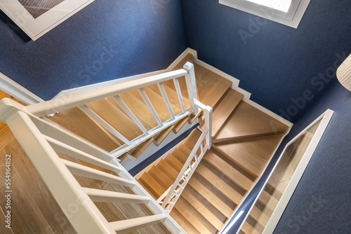 Top view of bright spiral staircase with polished wood parquet steps and matte white railings set against blue walls. Daylight streaming in from window illuminates spiral wooden staircase.