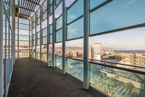 Long spacious terrace in high-rise building with fence and roof made of metal beams of strong impact-resistant glass protected from direct sunlight. Floor of terrace is covered with wooden parquet.