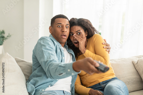 Scared shocked young african american woman and man with remote control switch channel on tv, watch horror film