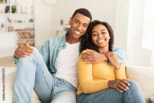 Cheerful handsome young african american man hugging his wife, resting on sofa, enjoy weekend © Prostock-studio