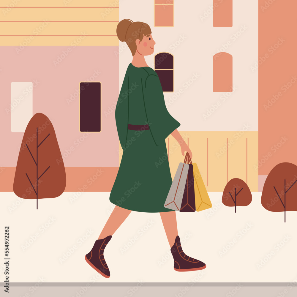 A woman in a green coat carries packages in her hand while walking down the street.