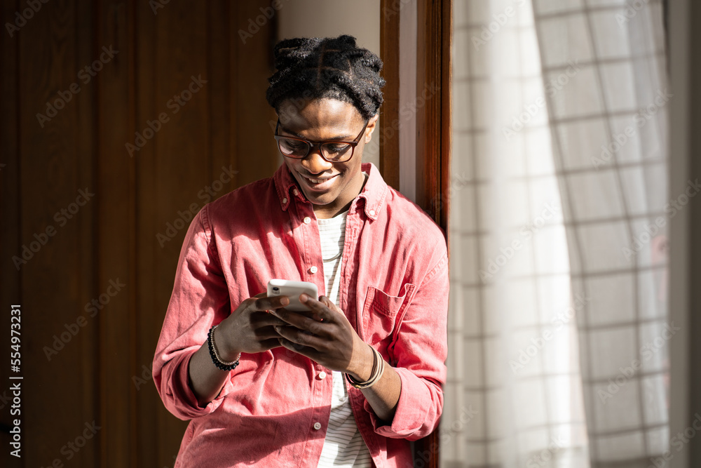 Happy smiling African guy holding cellphone typing, chatting online, using dating app while standing by window in sunny room at home. Young joyful black man enjoying leisure time with smartphone - obrazy, fototapety, plakaty 