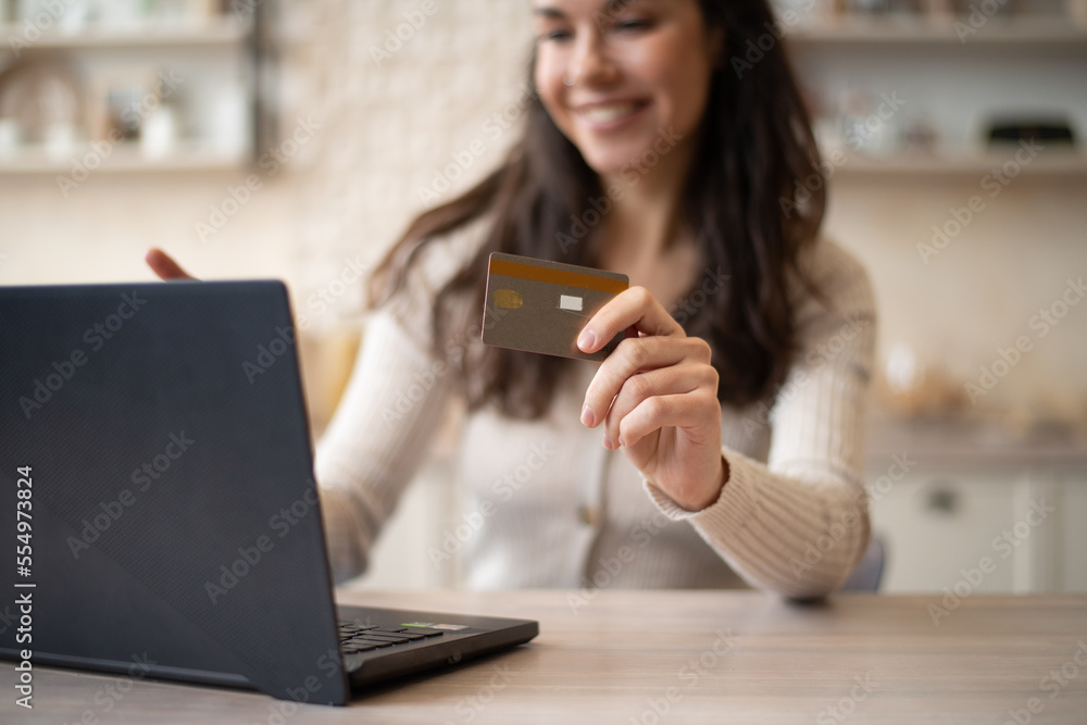 Happy lady with laptop and credit card buying purchases remotely, enjoying sales in kitchen interior, selective focus Happy lady with laptop and credit card buying purchases remotely, enjoying sales in kitchen interior, selective focus