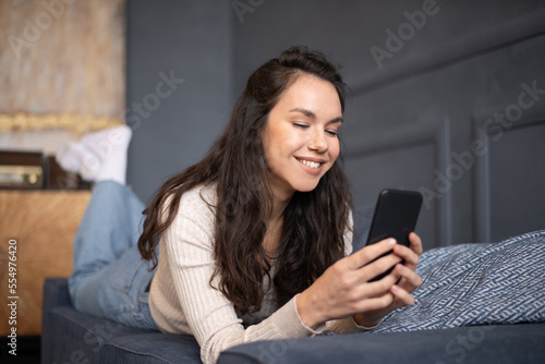 Happy lady lying on sofa and using smartphone, chatting with friends or surfing internet, copy space, home interior