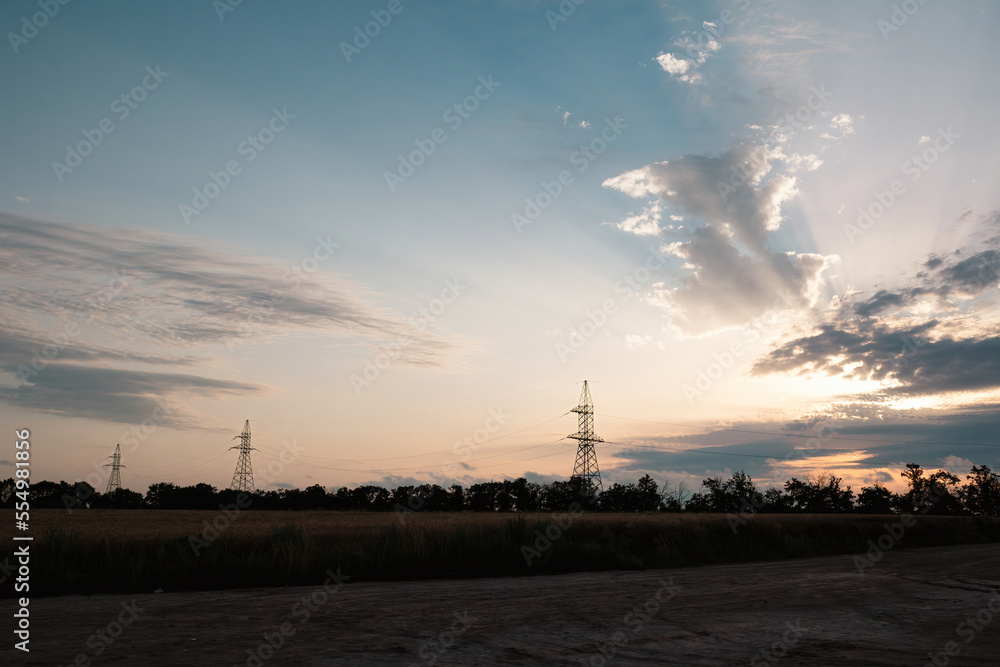 Sunset illuminates farmland with wheat fields and electrical towers silhouettes. Connected power transmission lines transfer renewable energy from windmill