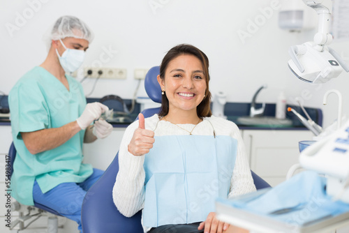 Happy cute latin woman sitting in dental chair after teeth cure giving thumb up