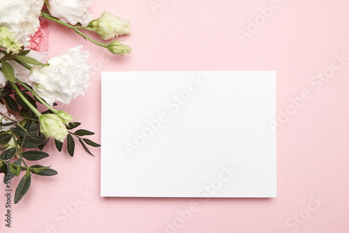 Holiday greeting card mockup with flowers on light pink background, top view, flat lay. White wedding invitation mockup and floral decor