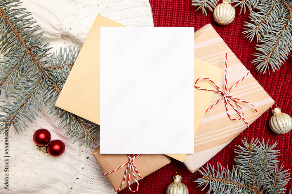 Obraz premium Christmas greeting card mockup with envelope, gift box, fir tree branches and holiday decorations on red knitted sweater background, top view, flat lay. White New Year card with winter decor