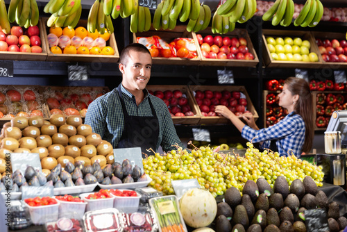 Friendly diligent cheerful man and woman laying out vegetables and fruits in shop photo
