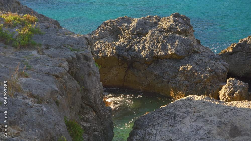 Calm clear green sea at shore with large rocks, sun shines to small waves, camera pans to side, typical scenery at one of many bays in Liapades, Greece