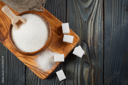 Wooden bowl with sugar and sugar cubes on a wooden plank on a dark wooden table, view from above