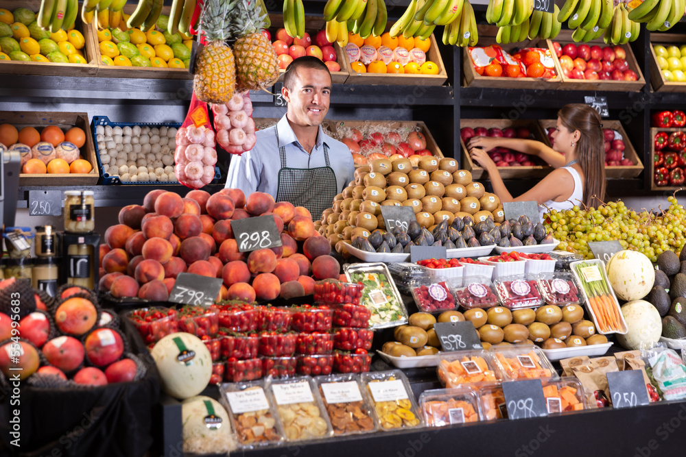 ..Portrait of young male worker in apron standing near fresh kiwi and fruits on the market