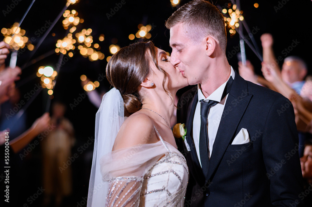 Portrait of loving couple kissing celebrating wedding day with guests friends on background. Night party of brides. Beautiful brunette woman in elegant wedding dress kissing groom. Sparkles. Happy. St...