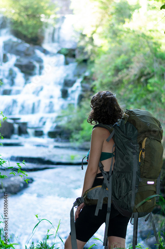 woman walking in nature with a camping backpack looks at the beautiful landscape where you can see a waterfall and a lot of green