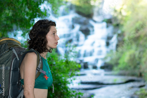 a woman hiking wears a camping backpack, looks at a waterfall surrounded by nature. landscape photo