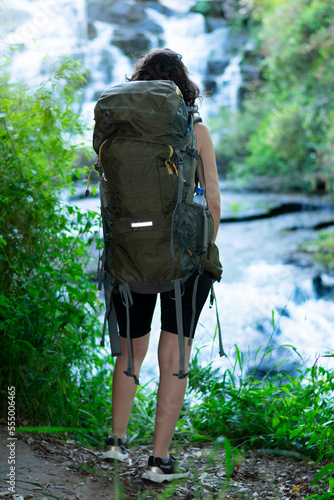 woman walking in nature with a camping backpack looks at the beautiful landscape where you can see a waterfall and a lot of green