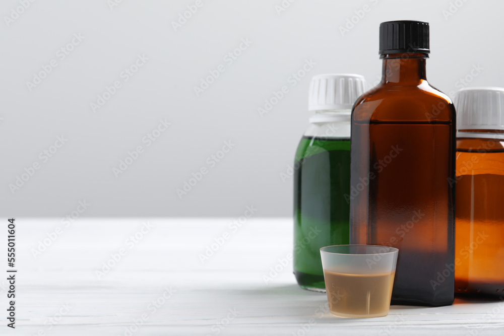 Bottles of syrup and measuring cup on white table against light grey background, space for text. Cold medicine Bottles of syrup and measuring cup on white table against light grey background, space for text. Cold medicine