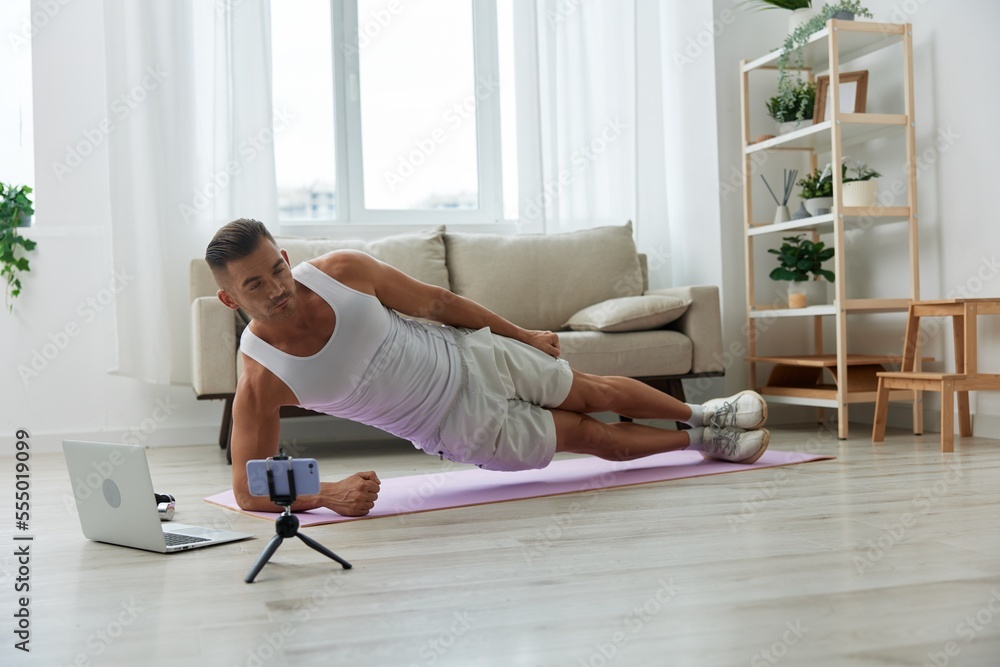 Man sports, watching tape of workout on phone and repeating exercises sports blogger with laptop training online, pumped up man fitness trainer works out at home, the concept of health and body beauty Man sports, watching tape of workout on phone and repeating exercises sports blogger with laptop training online, pumped up man fitness trainer works out at home, the concept of health and body beauty