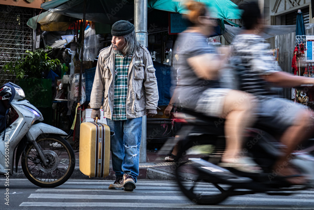 Obraz premium Asian poverty homeless man hold dragging large suitcase walk across the crosswalk in city. poor man live in slump no job, pity aged person problem from financial crisis unemployment without family