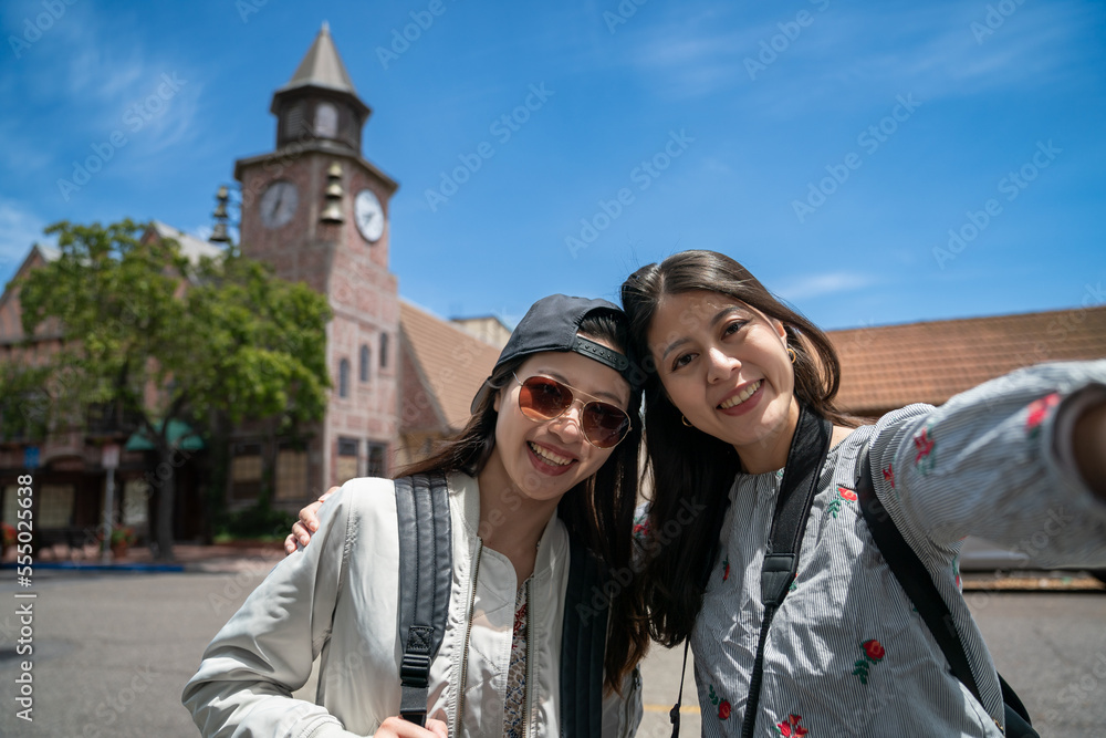 two happy asian Taiwanese girl visitors smiling at camera while taking selfie picture together with historical clock tower in the town of solvang under blue sky at background