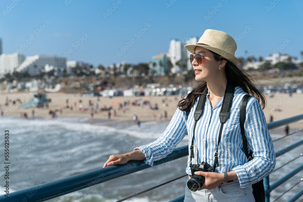 leisure asian Chinese woman traveler carrying camera looking over railing at santa monica beach. travel lifestyle and people on vacation in california usa