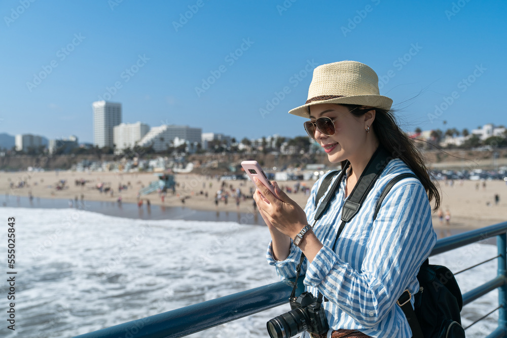 happy asian Japanese lady traveler on vacation searching guide on cellphone while relaxing near waterfront railing at beach