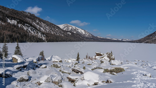 A chain of footprints is visible on the flat snow-covered surface of the frozen lake. Surrounding mountains against the blue sky. On the shore - a scattering of stones under a layer of snow, spruce. 