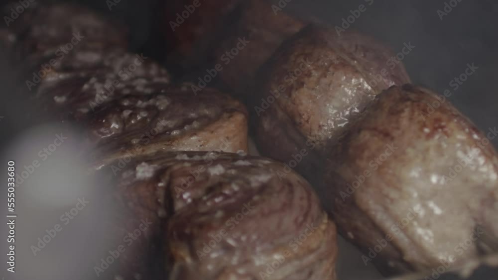 Close up, pork meat on skewers rotating, being grilled under smokey charcoal Close up, pork meat on skewers rotating, being grilled under smokey charcoal
