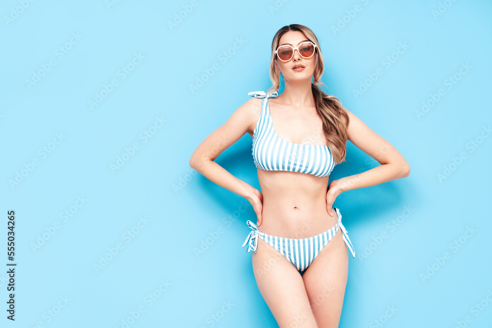 Fototapeta premium Young blond model in summer striped swimwear bathing suit. Sexy carefree woman having fun and going crazy. Female posing near blue wall in studio. Cheerful and happy. Perfect body. In sunglasses