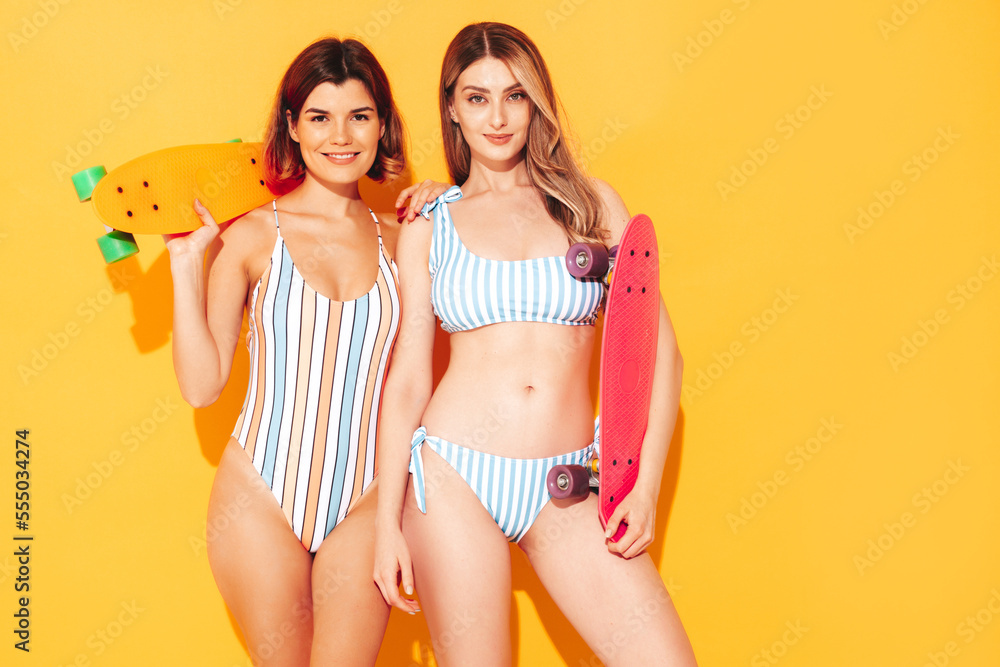 Two beautiful sexy smiling hipster women in summer swimwear bathing suits. Trendy models having fun in studio. Hot female isolated on yellow. Cheerful and happy. Holding penny skateboard