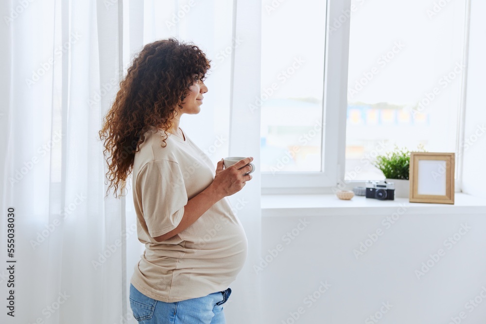 A pregnant woman stands by the window with a mug of warm water and tea and enjoys the view in a homemade t-shirt A pregnant woman stands by the window with a mug of warm water and tea and enjoys the view in a homemade t-shirt