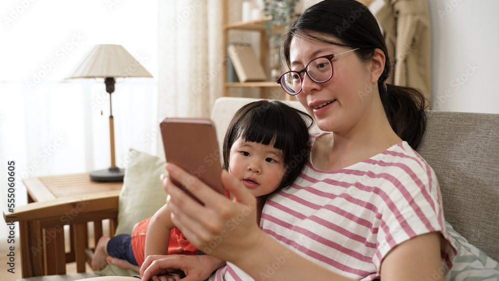 cute asian toddler baby girl snuggling with mother on sofa in living room while watching educational video for kid on the internet with phone together at home. cute asian toddler baby girl snuggling with mother on sofa in living room while watching educational video for kid on the internet with phone together at home.