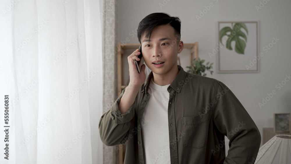 cheerful asian man receiving notification call on his smartphone while standing by the window at home. cheerful asian man receiving notification call on his smartphone while standing by the window at home.